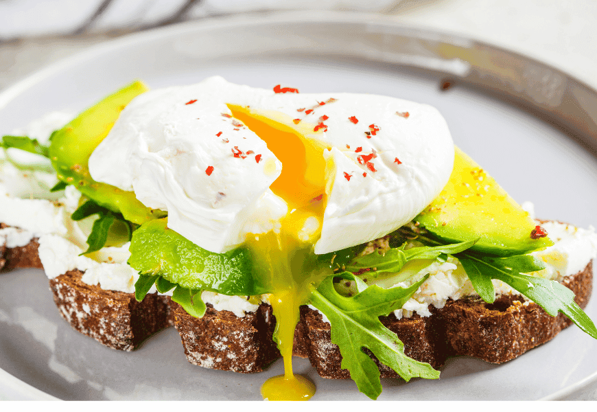 A slice of whole-grain bread topped with cream cheese, arugula, avocado slices, and a runny poached egg drizzled with tahini sauce and garnished with red pepper flakes, served on a gray plate.