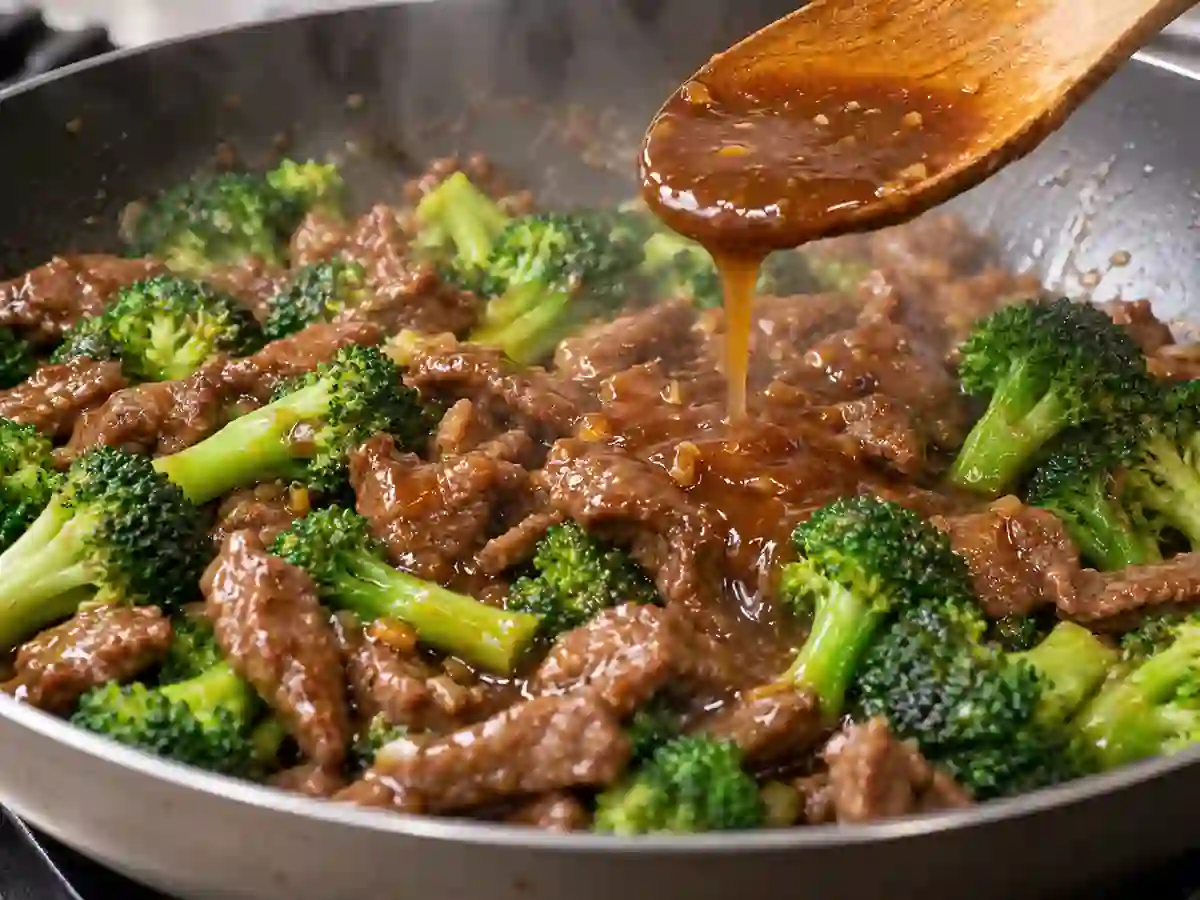 Glossy brown sauce coating a wooden spoon above beef and broccoli in the skillet.