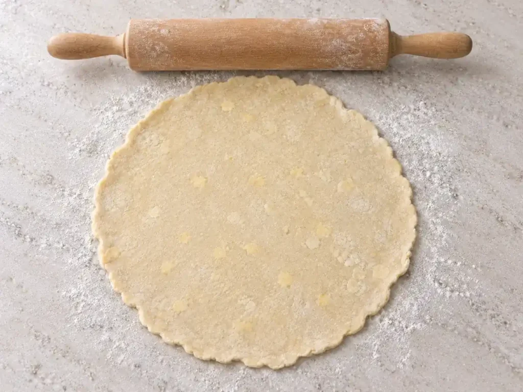 A thin round of raw pastry on a lightly floured counter with a wooden rolling pin