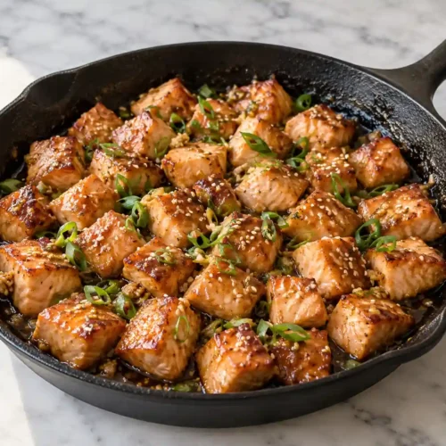 Honey garlic salmon bites in a black skillet with sesame seeds and sliced green onions.